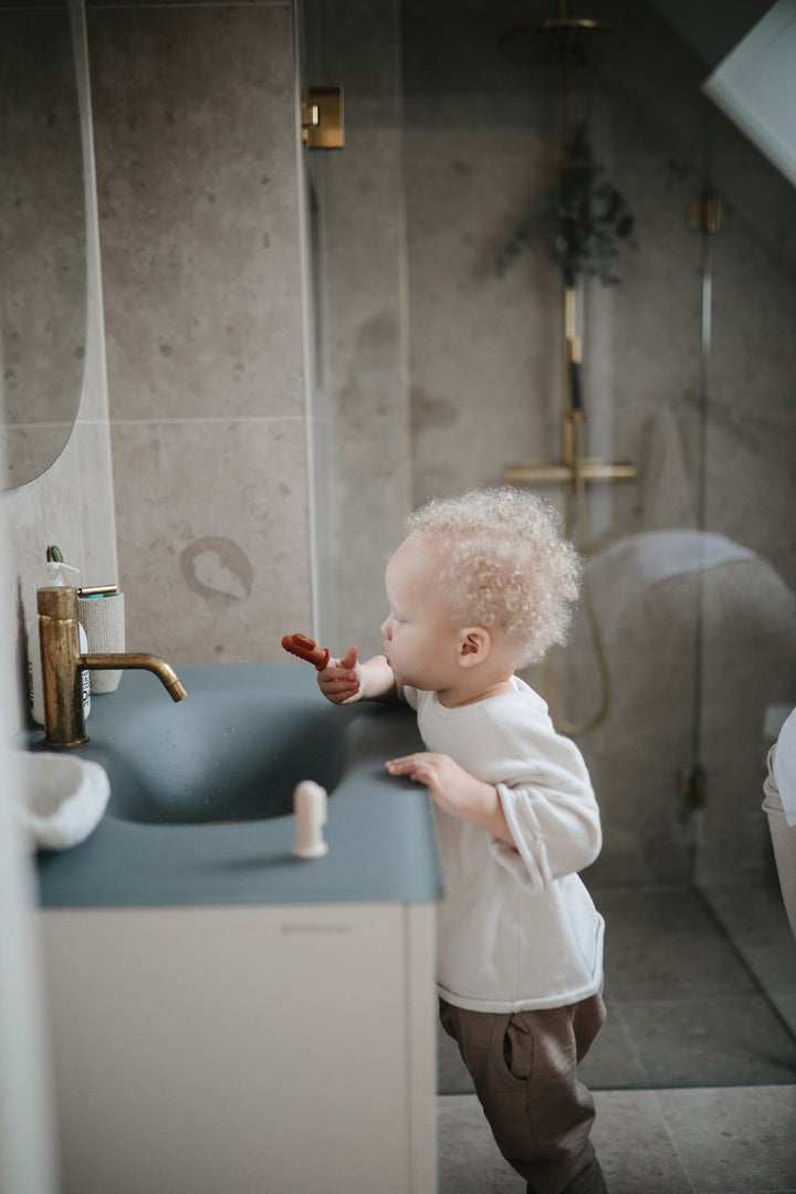 Blonde toddler boy brushing teeth with finger toothbrush at bathroom sink