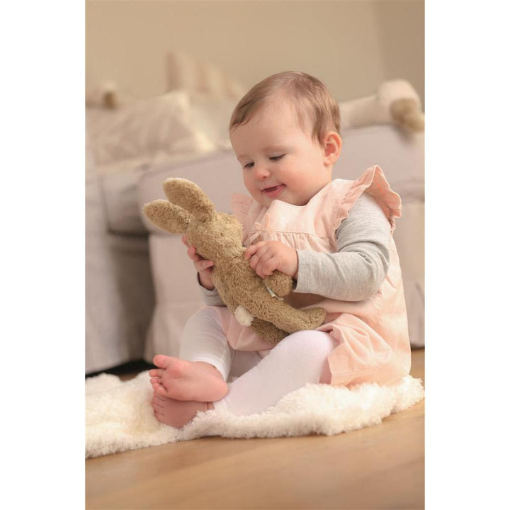Light brown soft plush toy rabbit bunny held and looked at by baby girl in pink dress sitting on floor