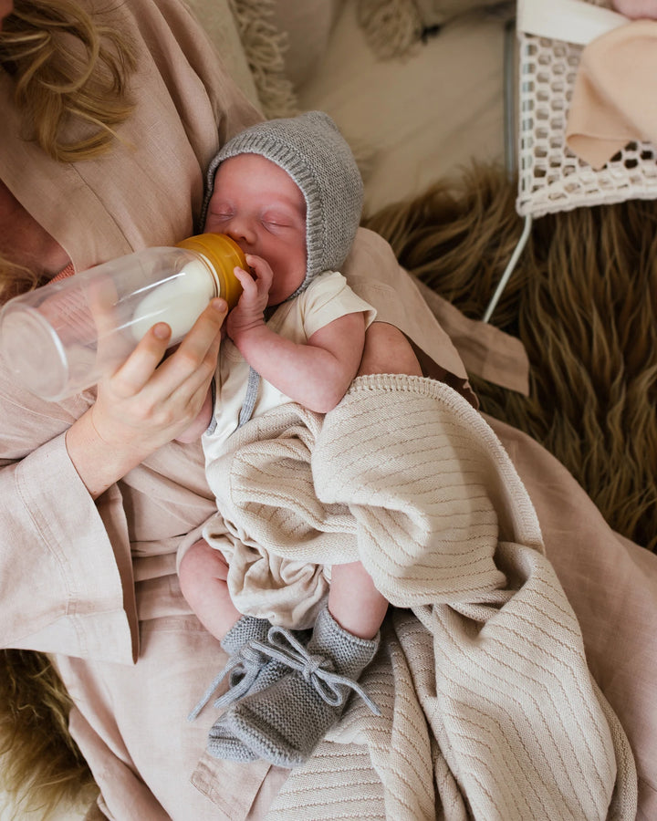 Newborn being bottle fed wearing grey Hvid bonnet and matching booties and Hvid Knitwear organic merino wool baby kids blanket Akira in off-white white natural ivory neutral