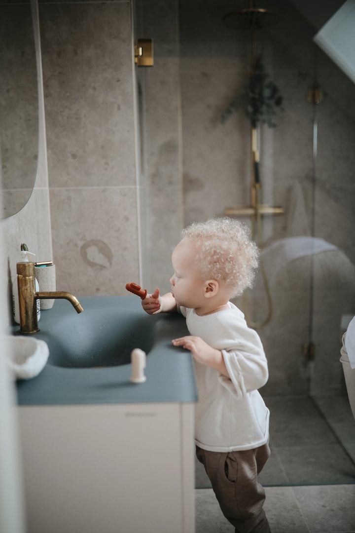 Blonde toddler boy brushing teeth with finger toothbrush at bathroom sink 