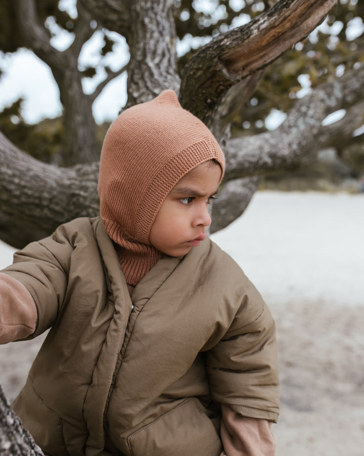 Toddler boy sitting o tree at beach wearing Hvid knitwear merino wool balaclava beanie and puffy winter jacket 