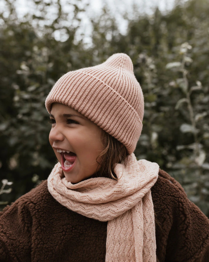 Girl wearing Hvid Knitwear Merino Wool Rib Winter Beanie Hat Fonzie for Kids and Toddlers in rose peach pink apricot and brown teddy jacket  standing outside in from of green bush