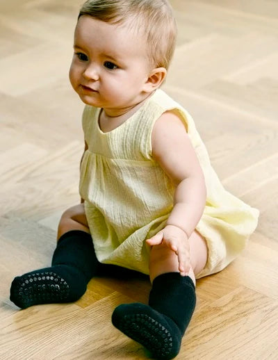 Baby sitting on floor wearing GoBabyGo cotton Terry non-slip socks wit rubber pads in dark grey melange and yellow sleeveless dress