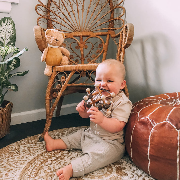 Baby boy sitting on floor holding and mouthing two tone wooden rattle toy in nursery in fornt of wicker chair with small teddy sitting on it 