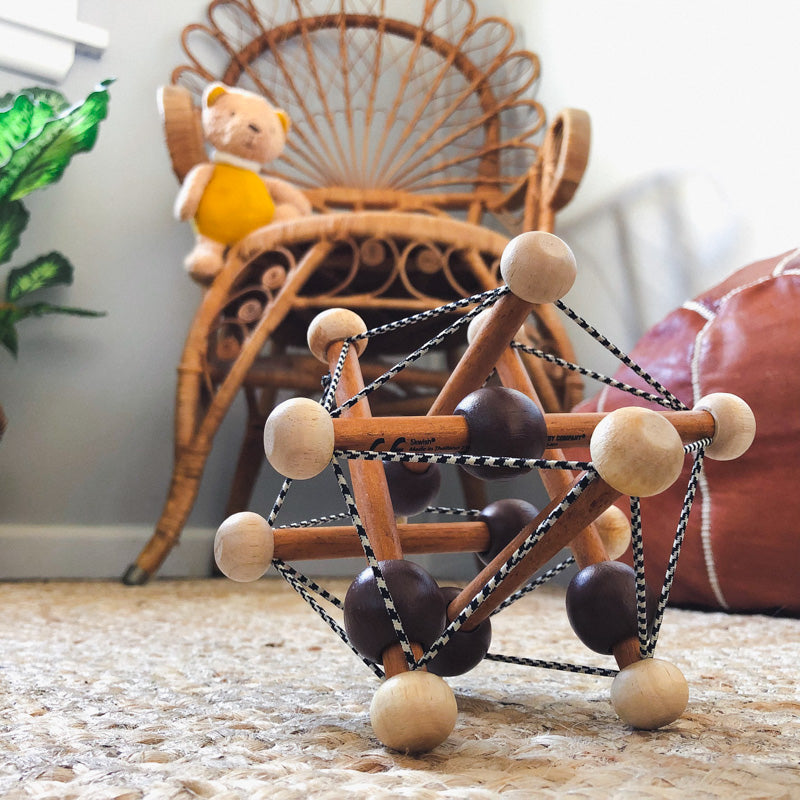 Two tone wooden rattle teether toy with strings lying on carpet floor in fornt of wicker chair with teddy bear on it, in nursery baby rom 