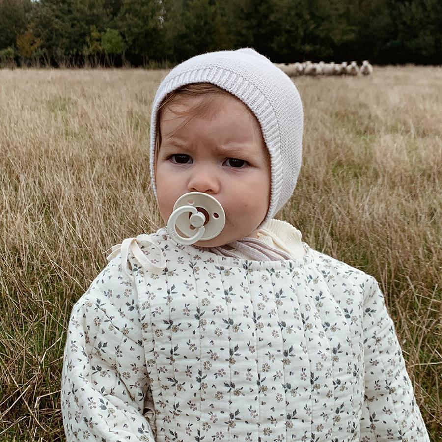 Baby toddler wearing white off-white natural ivory knitted wool bonnet sitting in field, white dummy in mouth